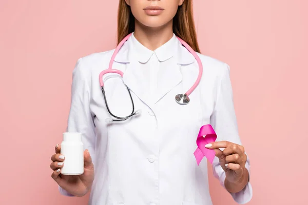 Vista recortada del médico sosteniendo frasco con pastillas y símbolo de conciencia de cáncer de mama aislado en rosa - foto de stock