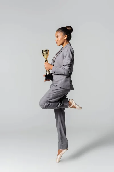Вид збоку афроамериканської бізнес-леді, що виконує балет із золотою чашкою ізольовано на сірому — стокове фото