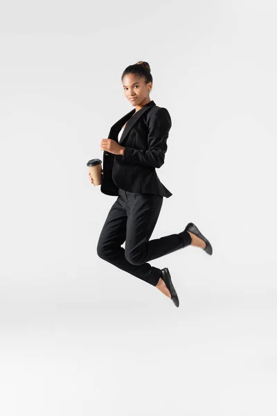Mujer de negocios afroamericana realizando ballet con taza de papel saltando aislado en gris - foto de stock