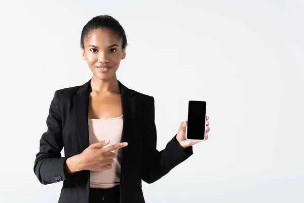 Улыбающаяся африканская американская бизнесвумен, указывающая на смартфон, изолированный на сером — стоковое фото