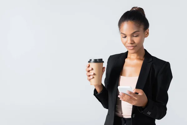 Mujer de negocios afroamericana con taza de papel usando teléfono inteligente aislado en gris - foto de stock