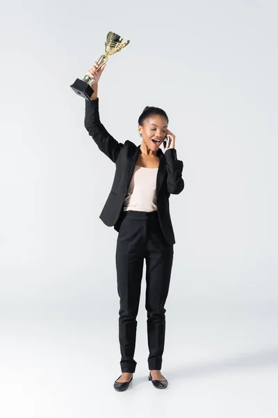 Happy african american businesswoman with golden cup talking on smartphone isolated on grey — Stock Photo