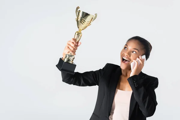 Feliz mujer de negocios afroamericana con copa de oro hablando en el teléfono inteligente aislado en gris - foto de stock