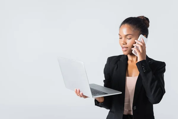 Mujer de negocios afroamericana con portátil hablando en teléfono inteligente aislado en gris - foto de stock