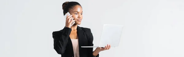 African american businesswoman with laptop talking on smartphone isolated on grey, banner — Stock Photo