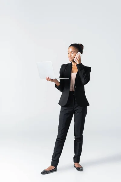 Femme d'affaires afro-américaine avec ordinateur portable parlant sur smartphone isolé sur gris — Photo de stock