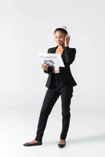 Donna d'affari afroamericana che ascolta musica in cuffia e legge giornali d'affari isolati su grigio — Foto stock