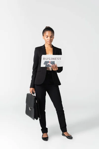 African american businesswoman with suitcase reading business newspaper isolated on grey — Stock Photo