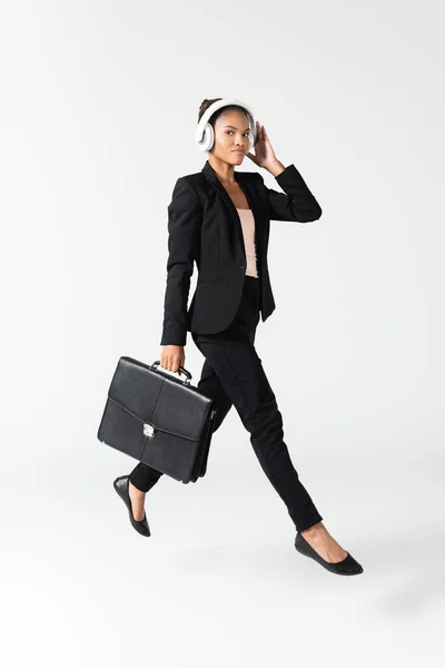 Африканская американская бизнесвумен в наушниках с чемоданом, изолированным на сером — стоковое фото