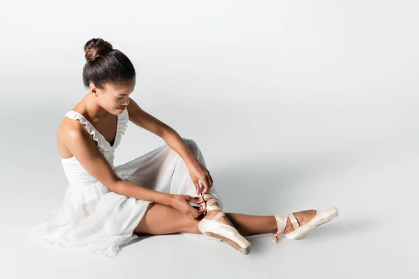 Graceful african american ballerina in dress adjusting pointe shoes on white background — Stock Photo