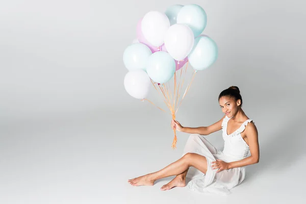 Descalza elegante bailarina afroamericana en vestido sentado con globos sobre fondo blanco - foto de stock