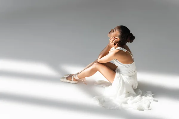 Graceful african american ballerina in dress sitting on floor on white background — Stock Photo
