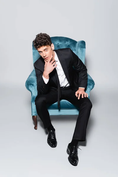 Элегантный мужчина в костюме сидит в велюровом кресле изолированный на сером — стоковое фото