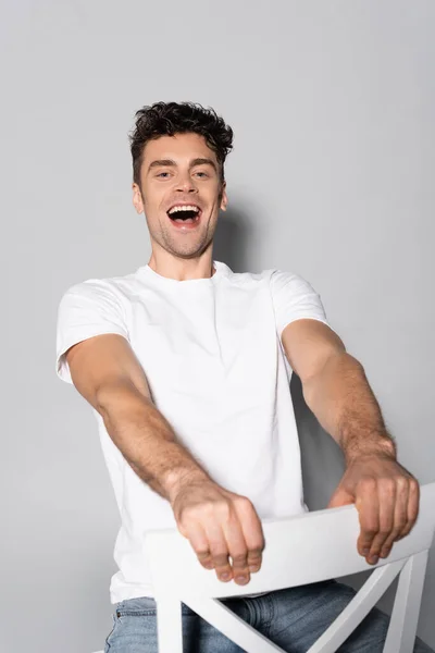 Jovem feliz em t-shirt branca na cadeira isolada no cinza — Fotografia de Stock