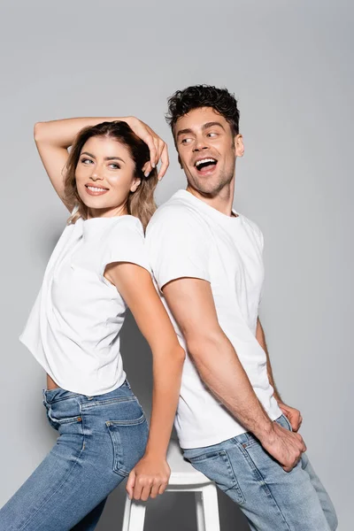 Sonriente pareja joven en camisetas blancas y jeans posando espalda con espalda aislados en gris - foto de stock