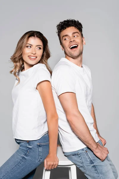 Улыбающаяся молодая пара в белых футболках и джинсах, позирующая спина к спине, изолированная на сером — стоковое фото