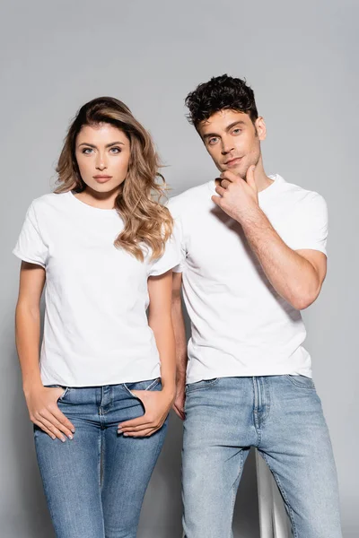 Jeune couple en t-shirts et jeans blancs posant isolé sur gris — Photo de stock