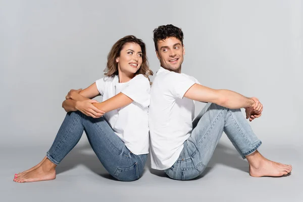 Giovane coppia in t-shirt bianche e jeans in posa schiena a schiena isolata su grigio — Foto stock