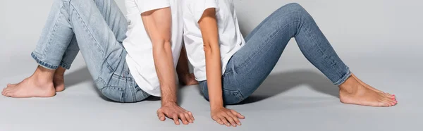 Vista recortada de pareja joven en camisetas blancas y jeans posando espalda con espalda aislados en gris, pancarta - foto de stock