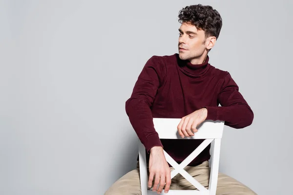 Young man in burgundy jumper posing on chair isolated on grey — Stock Photo