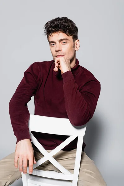 Young man in burgundy jumper posing on chair isolated on grey — Stock Photo