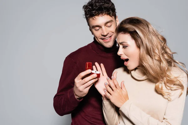 Jovem casual fazendo proposta de casamento para mulher chocada isolado em cinza — Fotografia de Stock