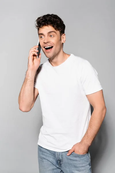 Jovem feliz em t-shirt branca falando no smartphone isolado em cinza — Fotografia de Stock