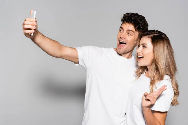 Feliz pareja joven casual en camisetas blancas tomando selfie en el teléfono inteligente aislado en gris - foto de stock