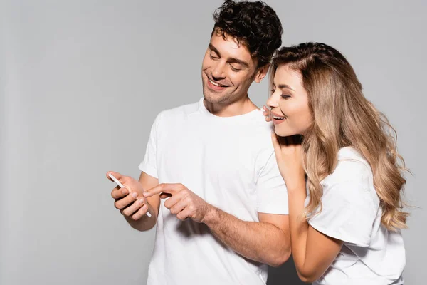 Feliz pareja joven casual en camisetas blancas con teléfono inteligente aislado en gris - foto de stock