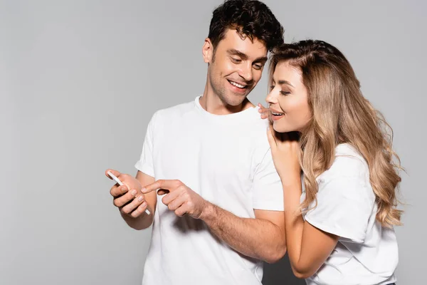 Feliz pareja joven casual en camisetas blancas con teléfono inteligente aislado en gris - foto de stock