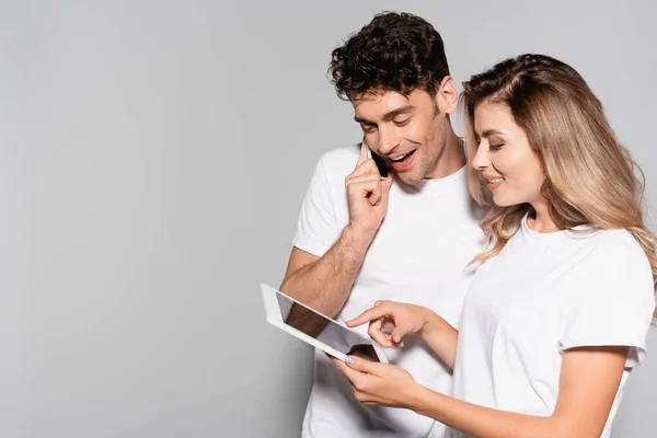 Feliz pareja joven casual en camisetas blancas con teléfono inteligente y tableta digital aislada en gris - foto de stock
