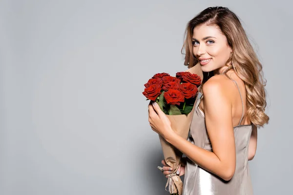 Donna elegante e sorridente in abito con bouquet di rose rosse isolate su grigio — Foto stock