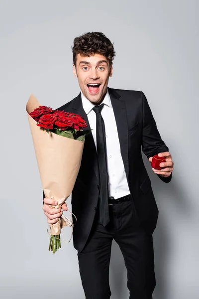 Uomo sorridente in abito elegante con bouquet di rose rosse e scatola anello isolato su grigio — Foto stock