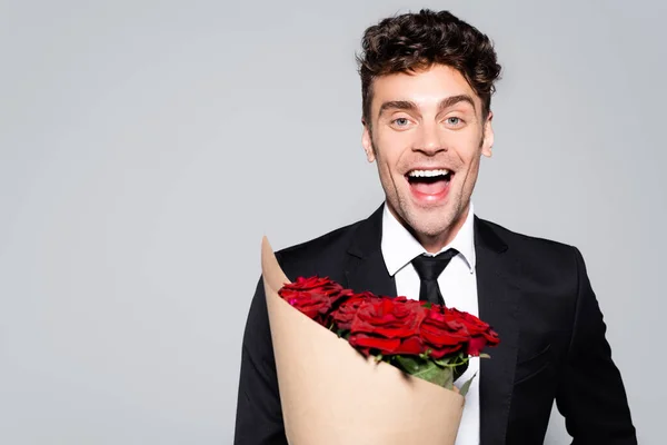 Happy man in elegant suit with bouquet of red roses isolated on grey — Stock Photo