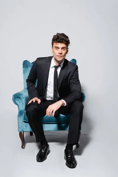 Elegante hombre de traje sentado en sillón de terciopelo aislado en gris - foto de stock