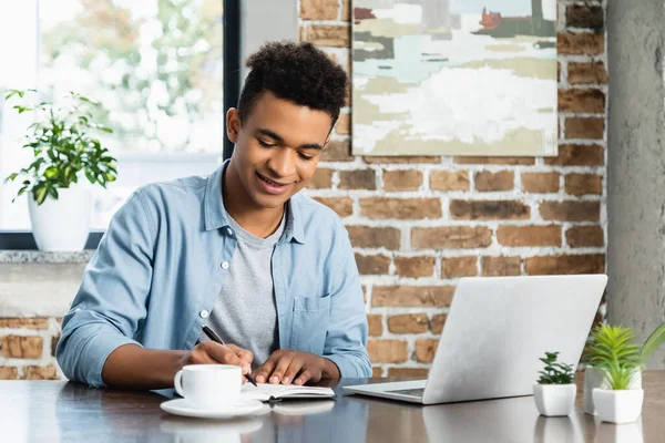 Щасливий афроамериканський чоловік пише в блокноті біля ноутбука і чашки на столі — стокове фото