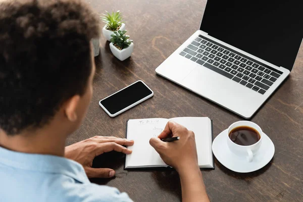 Hochwinkelige Ansicht eines afrikanisch-amerikanischen Mannes, der in einem Notizbuch in der Nähe von Geräten und einer Tasse Kaffee auf dem Schreibtisch schreibt — Stockfoto