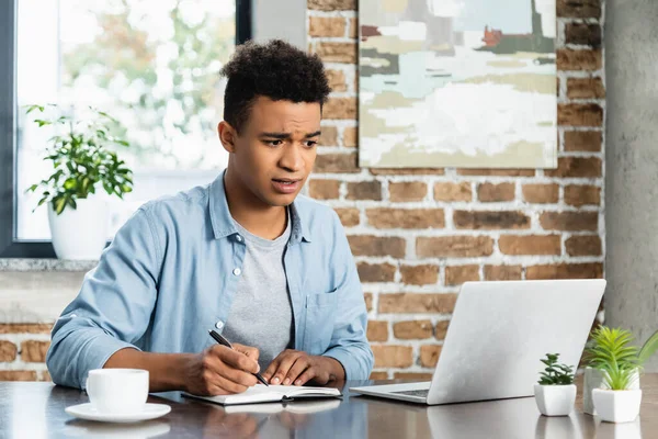 Hombre afroamericano sorprendido sosteniendo pluma cerca de portátil y mirando a la computadora portátil - foto de stock