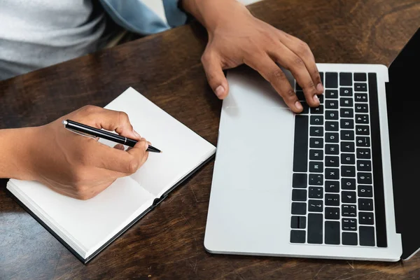 Vista ritagliata dell'uomo africano americano che tiene la penna vicino al notebook e usando il computer portatile — Foto stock