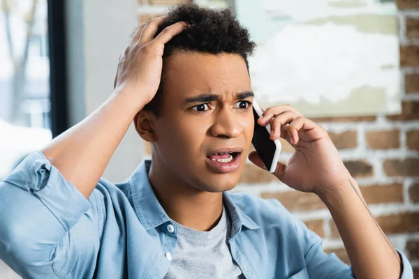 Chocado homem americano africano falando no smartphone e ajustando o cabelo encaracolado — Fotografia de Stock