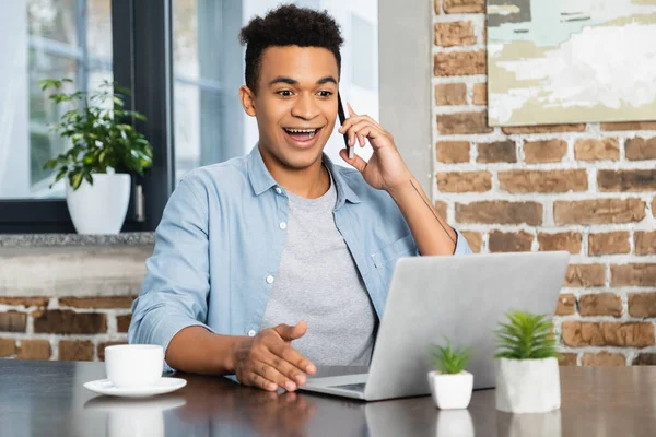 Asombrado hombre afroamericano hablando en teléfono inteligente cerca de la computadora portátil en el escritorio - foto de stock