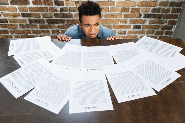 Choqué homme afro-américain regardant des documents avec des lettres contractuelles sur le bureau — Photo de stock