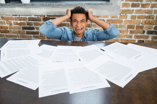 Homme afro-américain hystérique tirant les cheveux près des documents avec lettrage contractuel sur le bureau — Photo de stock
