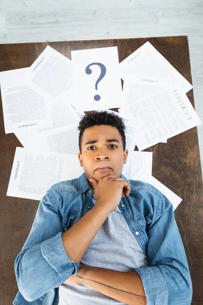 Vista superior del hombre afroamericano pensativo mirando la cámara cerca de documentos con letras de contrato en el escritorio - foto de stock