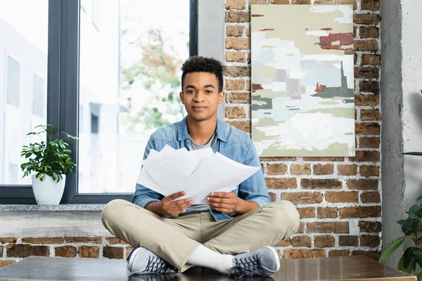 African american man sitting with crossed legs on desk and holding documents — Stock Photo