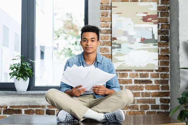 Homme afro-américain assis avec les jambes croisées sur le bureau et tenant des documents — Photo de stock