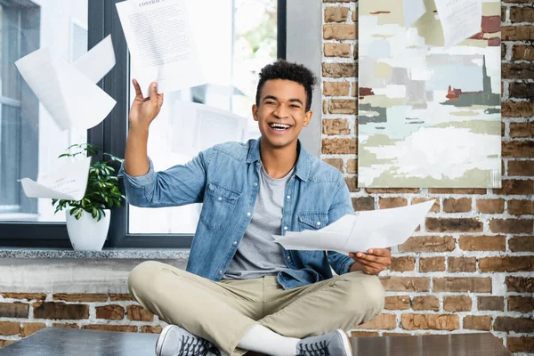 Glücklicher afrikanisch-amerikanischer Mann sitzt mit gekreuzten Beinen auf dem Schreibtisch und wirft Luftdokumente ein — Stockfoto