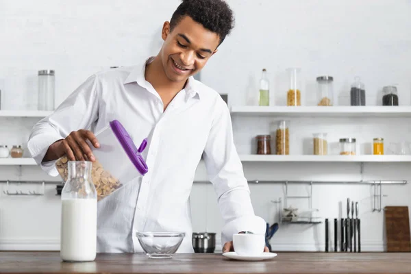 Hombre afroamericano feliz sosteniendo contenedor con hojuelas de maíz cerca de tazón y bebidas en la mesa - foto de stock