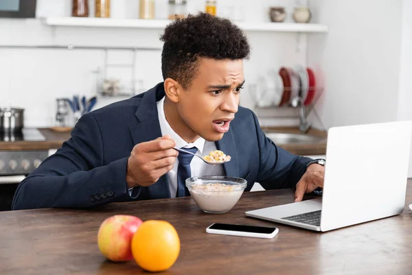 Sorprendido afroamericano freelancer en traje usando el ordenador portátil mientras desayunaba - foto de stock