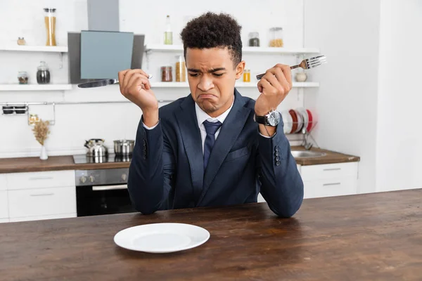 Sad african american man holding cutlery and looking at empty plate — Stock Photo